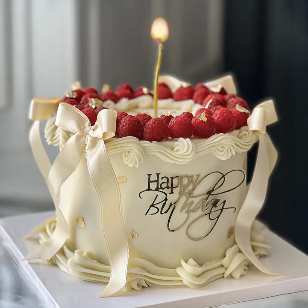 Krem rengi, ahududu, altın kurdeleli, Happy Birthday yazılı ve yanan mumlu doğum günü pastası.