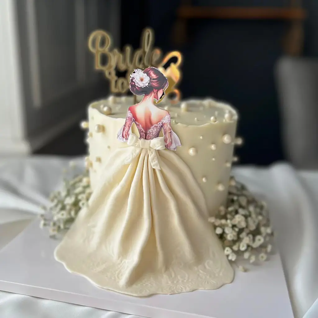 Krem rengi incili pasta üzerinde altın Bride to be yazısı ve dantel detaylı gelin elbiseli figür.